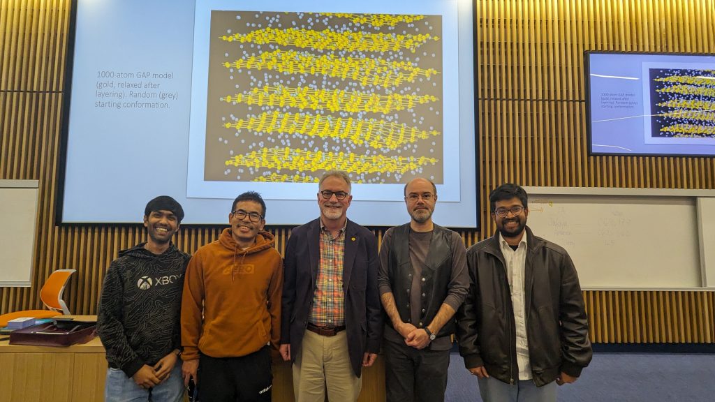 Professors and students standing together in a large classroom against a background of a projector screen.
